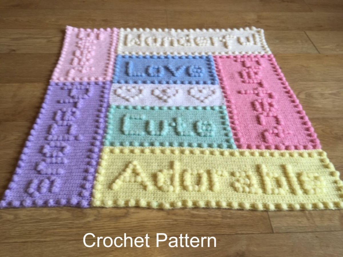 Multi colored crochet blanket featuring rectangles with words stitched into it and a small trim of bobbles laid on a wooden background.