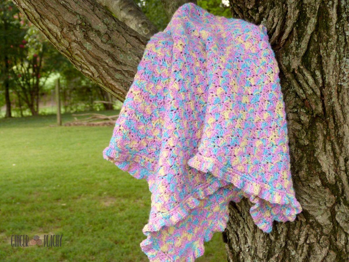  Pink, yellow, and blue crochet blanket with slight scalloped trim hanging from a tree with grass in the background. 
