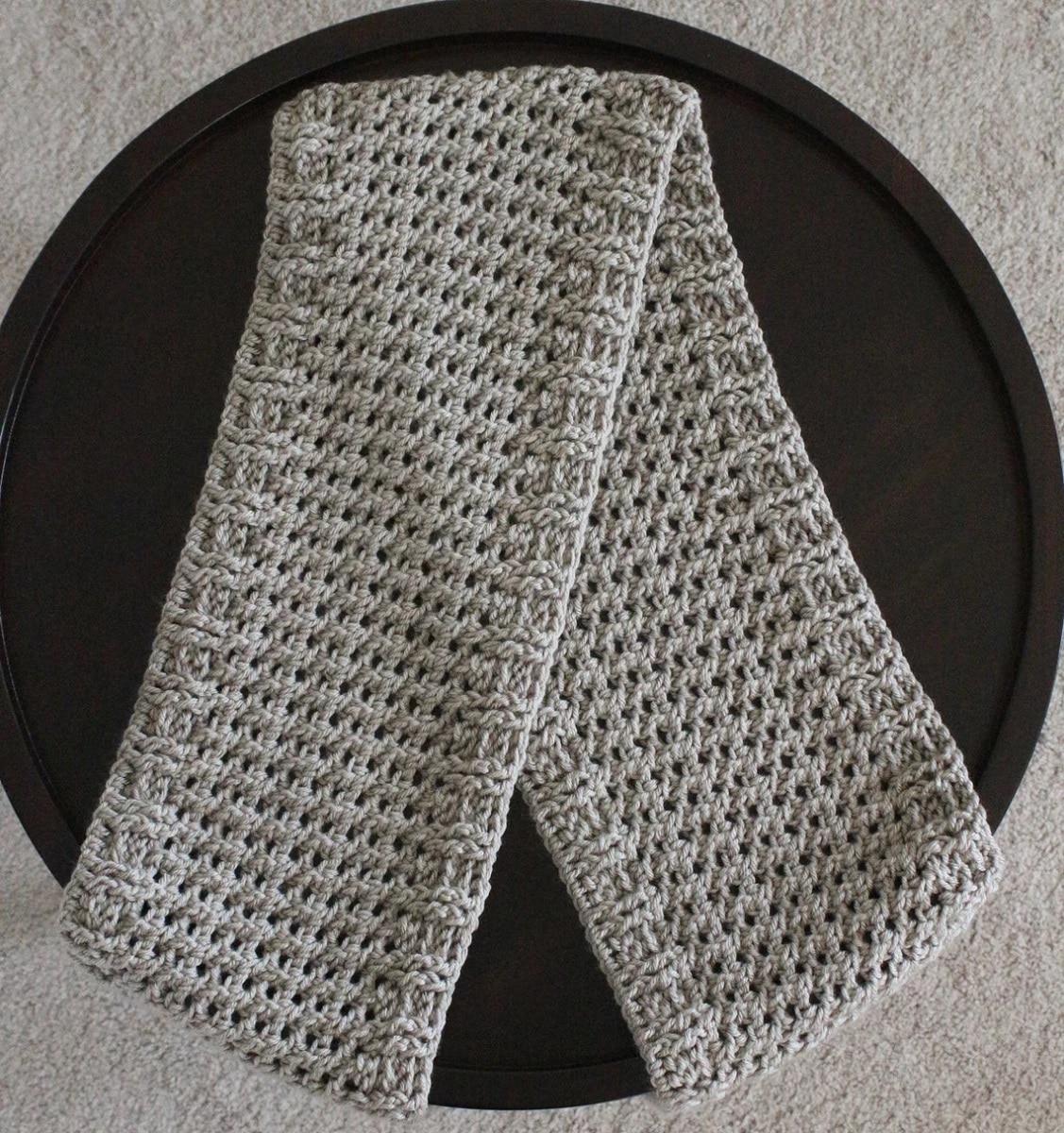 A light gray crochet scarf with a tight waffle stitch in the middle folded over on a black table.