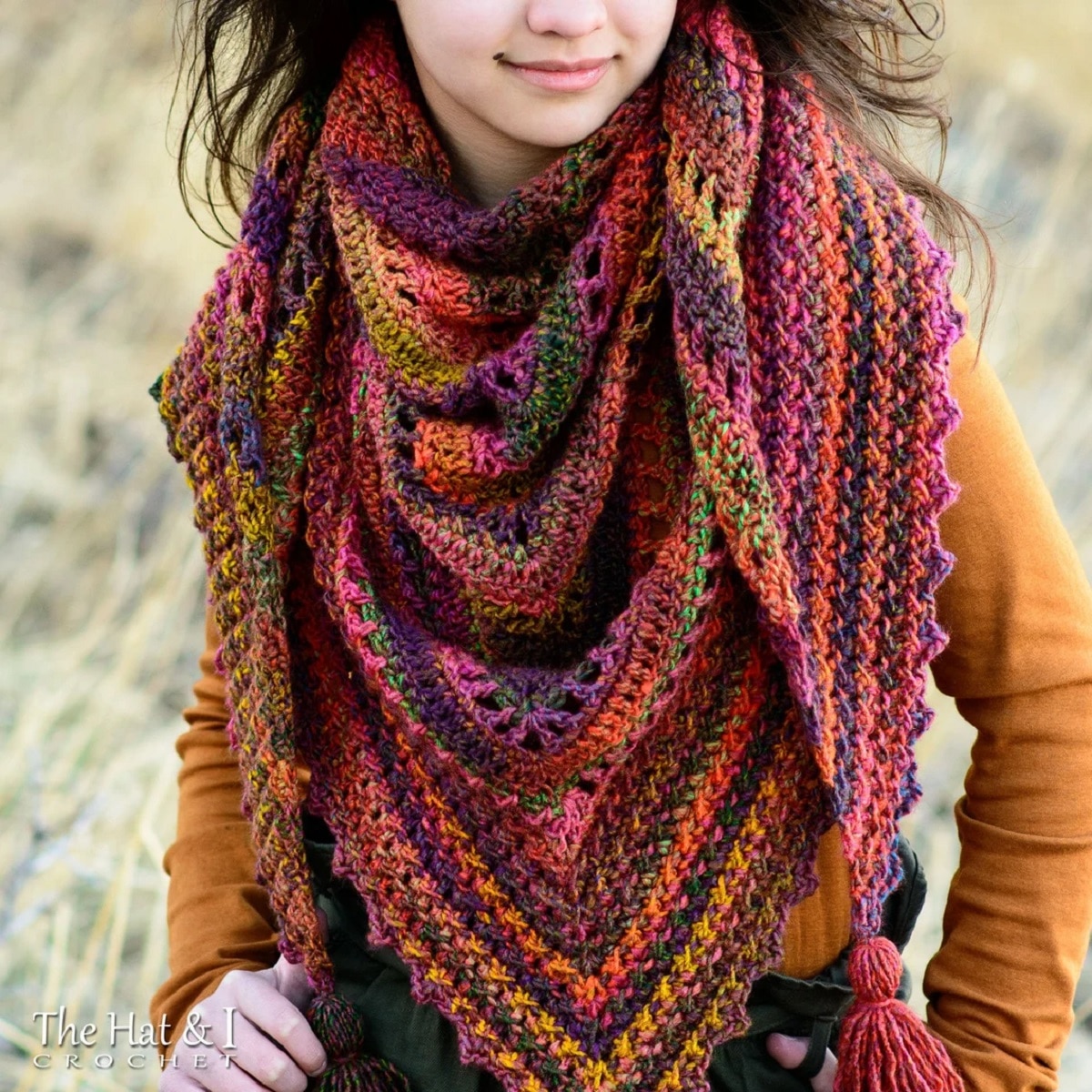 Brunette woman smiling with a pink, purple, green, yellow, and brown striped crochet triangle scarf around her neck.