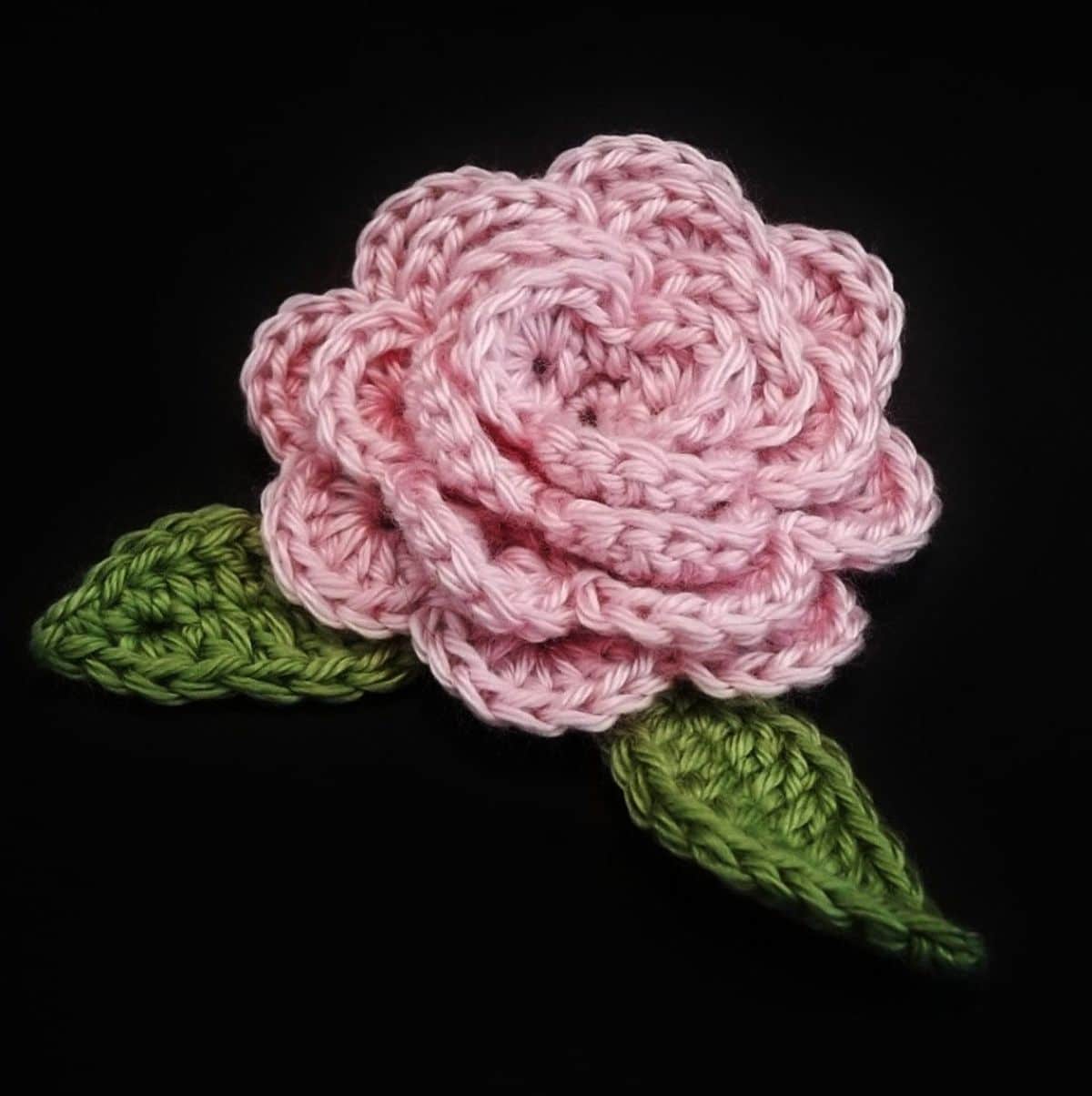 A light pink crochet rose with two green leaves either side on a black background.