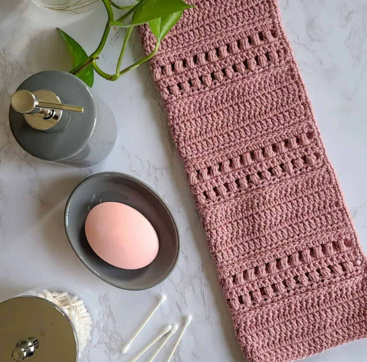 A pink crochet towel on a marble countertop next to a soap dish, some Q-tips, and a soap dispenser.