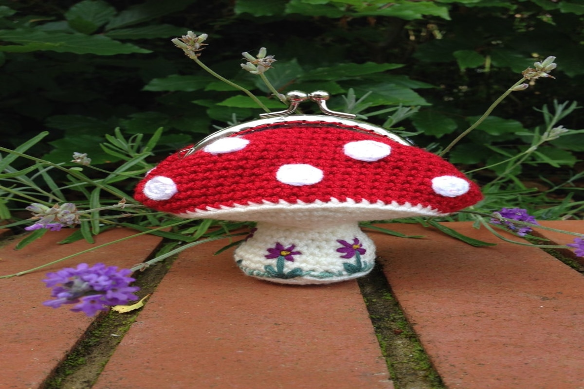 White crochet toadstool coin purse with purple flowers along the base and a red and white spotty top with the opening to the purse attached.