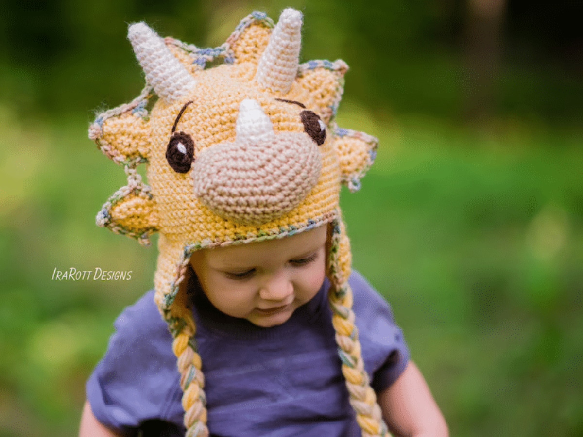 Baby sat outside wearing a light orange Triceratop hat with light brown horns, nose, and orange and brown braids on either side.