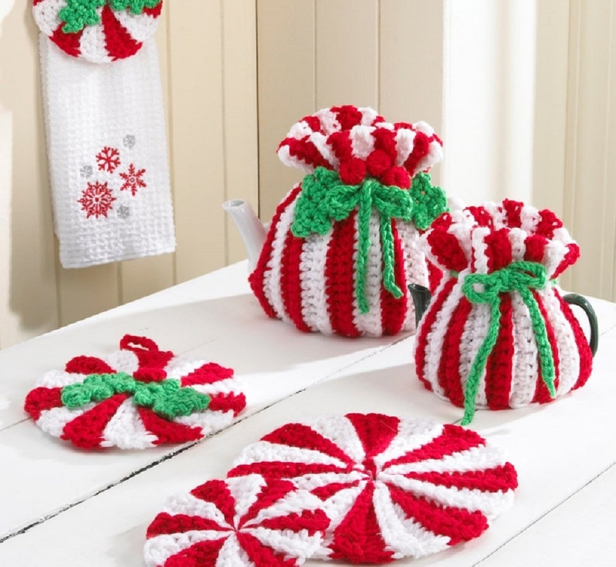 Red and white peppermint style coasters next to a tea cozy and milk jug cover in the same style with a green bow around them. 