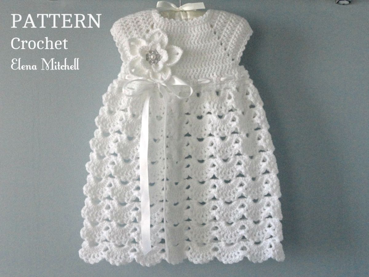 White crochet baby dress with short sleeves, a white flower on the left with satin rope hanging down and a full skirt.