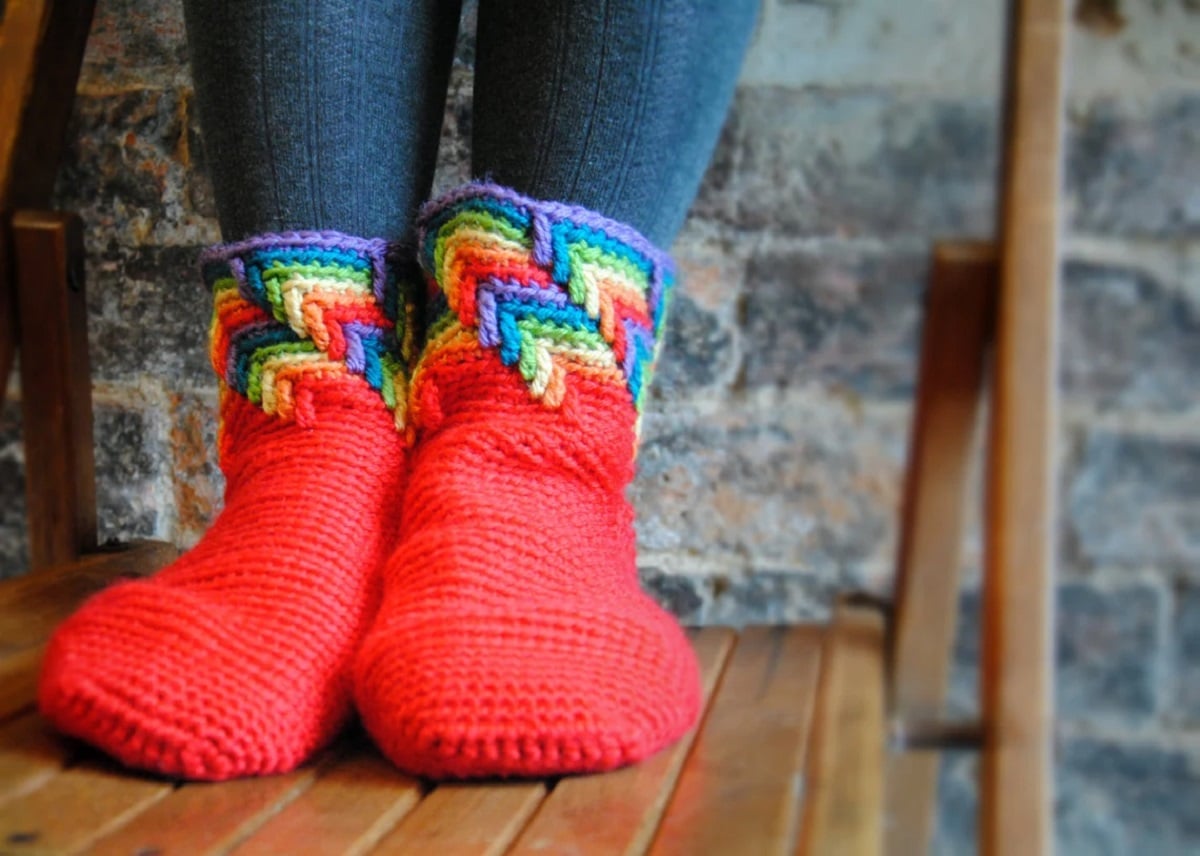 Feet wearing thick red crochet socks with a rainbow trail of tears pattern at the top around the ankle and lower calf.