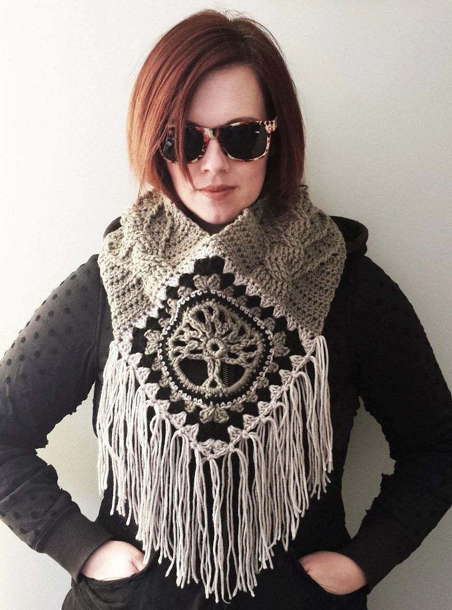 Beige, green, black, and white crochet cowl with a tree of life in the centre and white tassels dangling on the bottom.