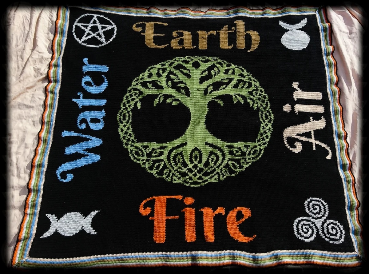 Black crochet blanket with a green tree of life in the center and the four elements listed on the sides with their pagan symbols next to them.