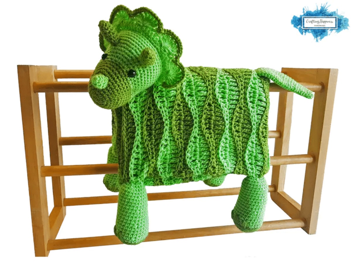 Green crochet baby blanket with a stuffed Triceratop head with gray horns and four green stuffed legs draped over a wooden crib.