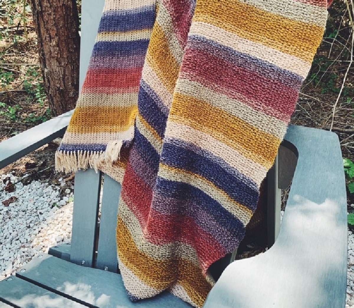 A white, yellow, blue, purple, and pink horizontal striped crochet Afghan hanging over a blue chair and some snow.