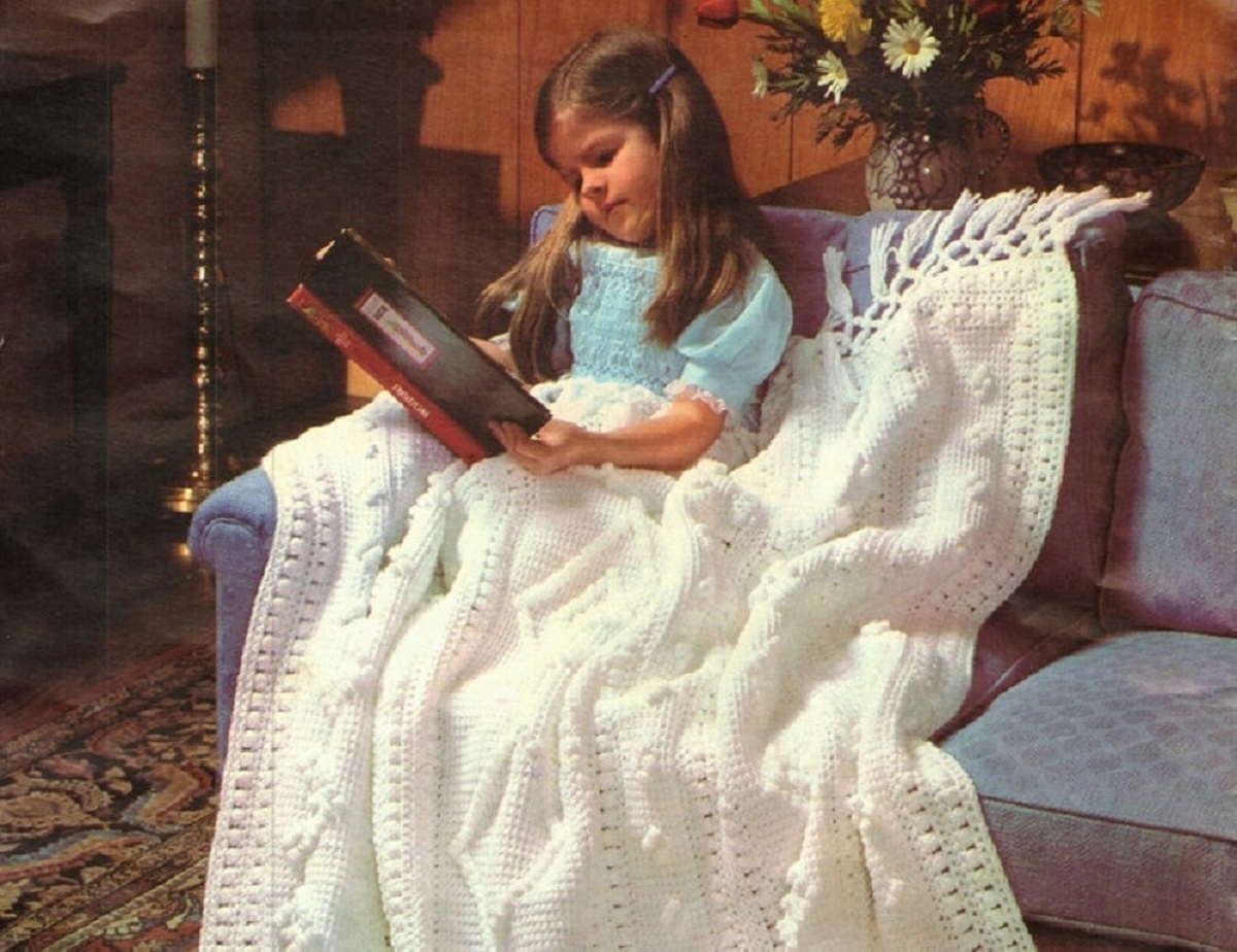 Young girl in a blue dress reading a book under a large white crochet blanket with tassels along the top.