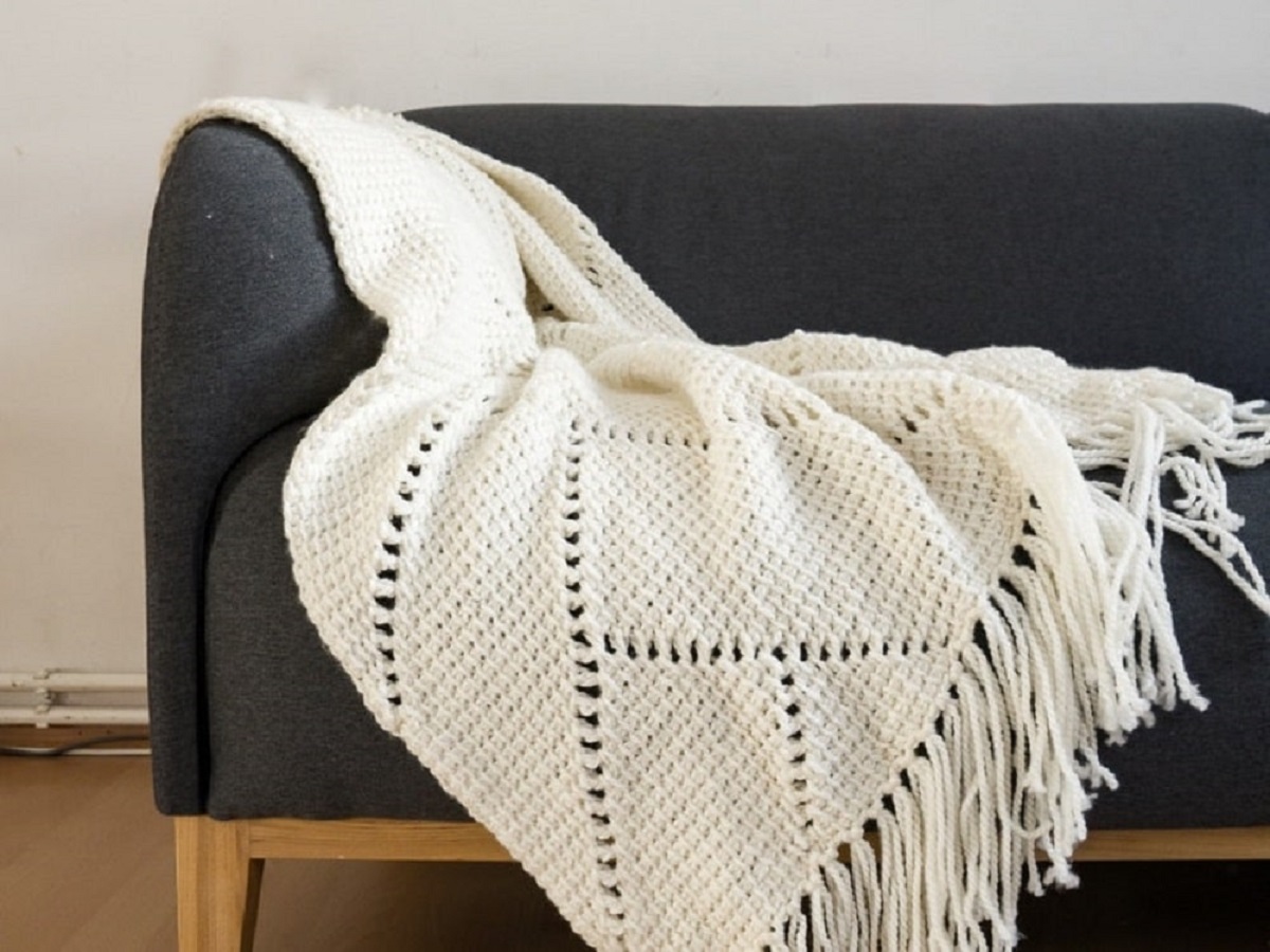White herringbone style crochet Afghan with long tassels along the bottom draped over the arm of a navy couch.