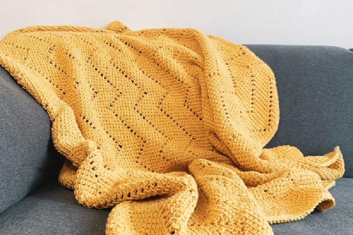 A yellow crochet Afghan with vertical zig zags bunched up and folded on a gray couch.