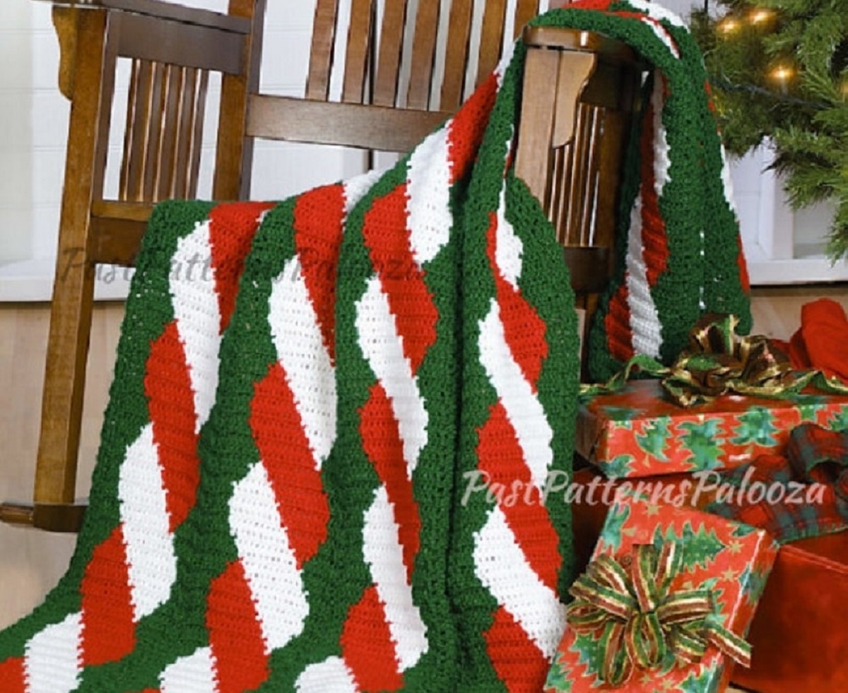 Green crochet blanket with red and white candy cane stripes down the center draped over a wooden chair next to wrapped gifts.