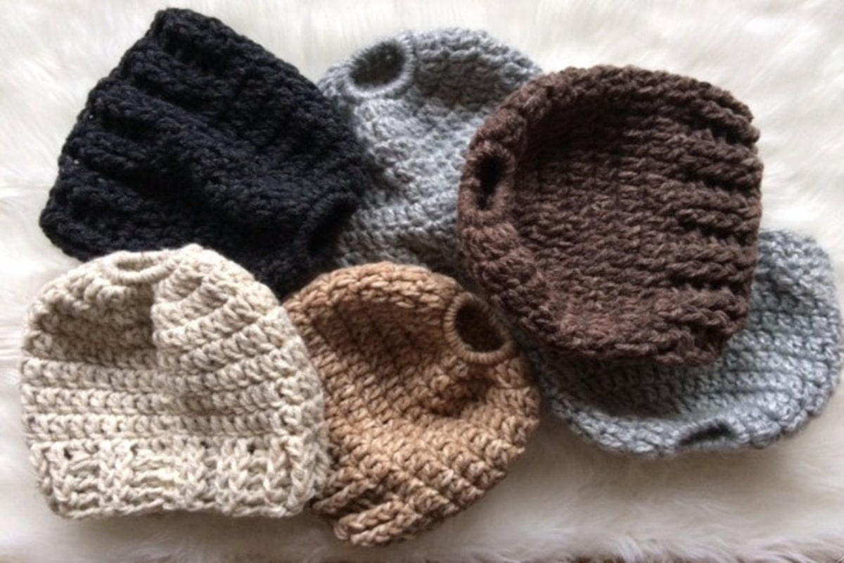 A small pile of cream, light brown, brown, gray, and black crochet hats with holes at the top on a cream blanket.