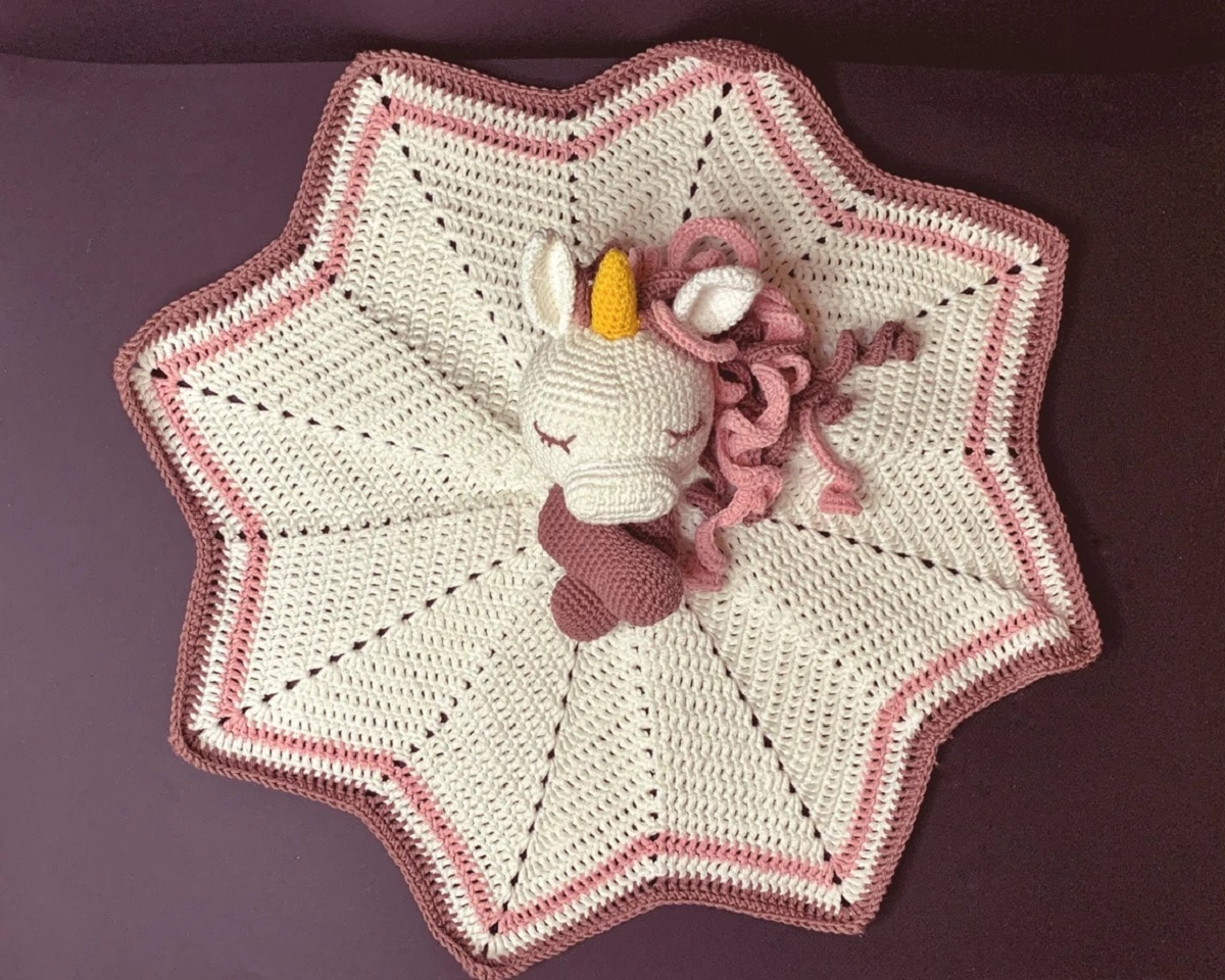 A white crochet star shaped blanket with a pink trim and a stuffed unicorn head, horn, mane, and arms attached to the center of the blanket.