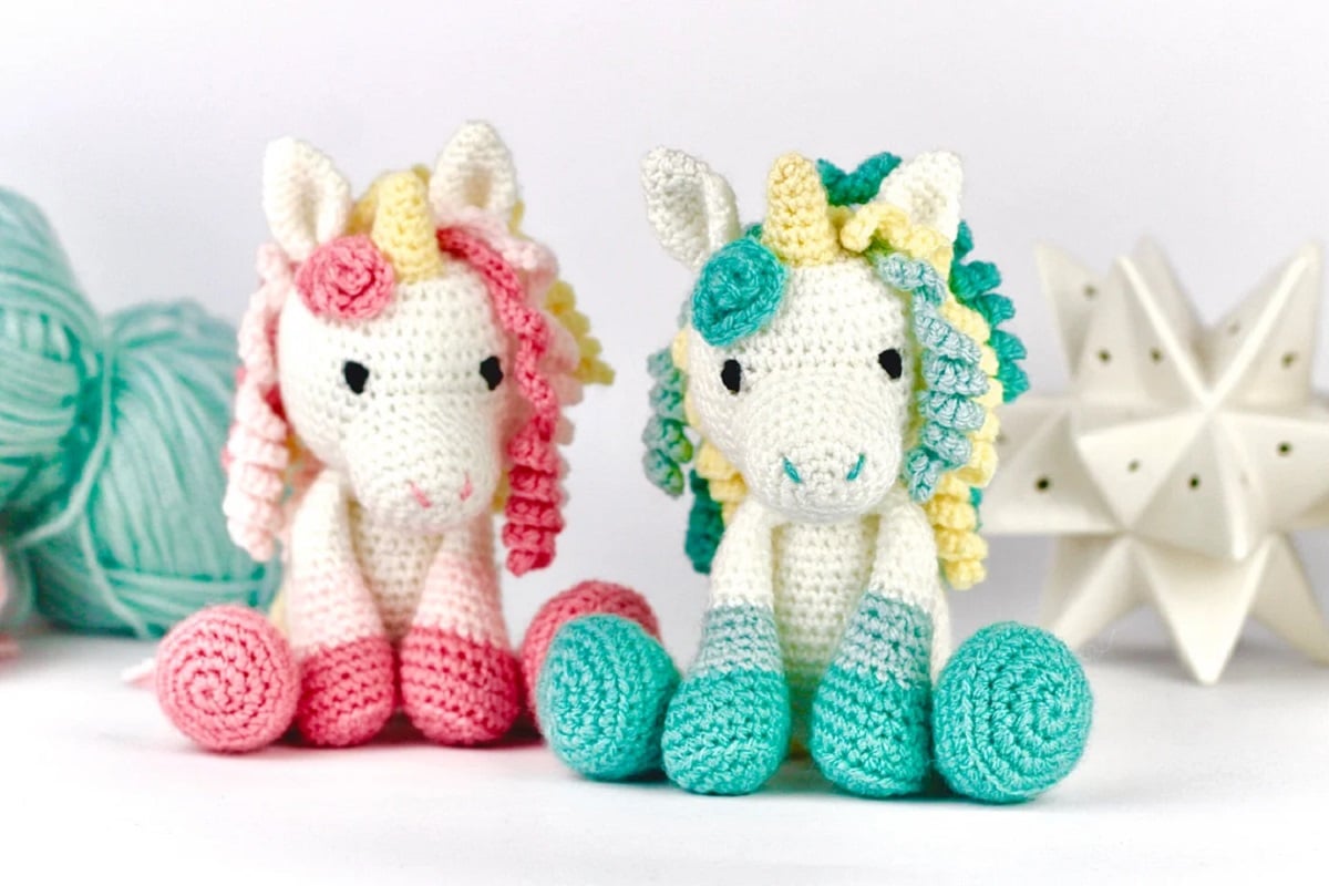 A white crochet unicorn with a pink mane, feet, and yellow horn, sitting next to a white unicorn with the same features in blue.