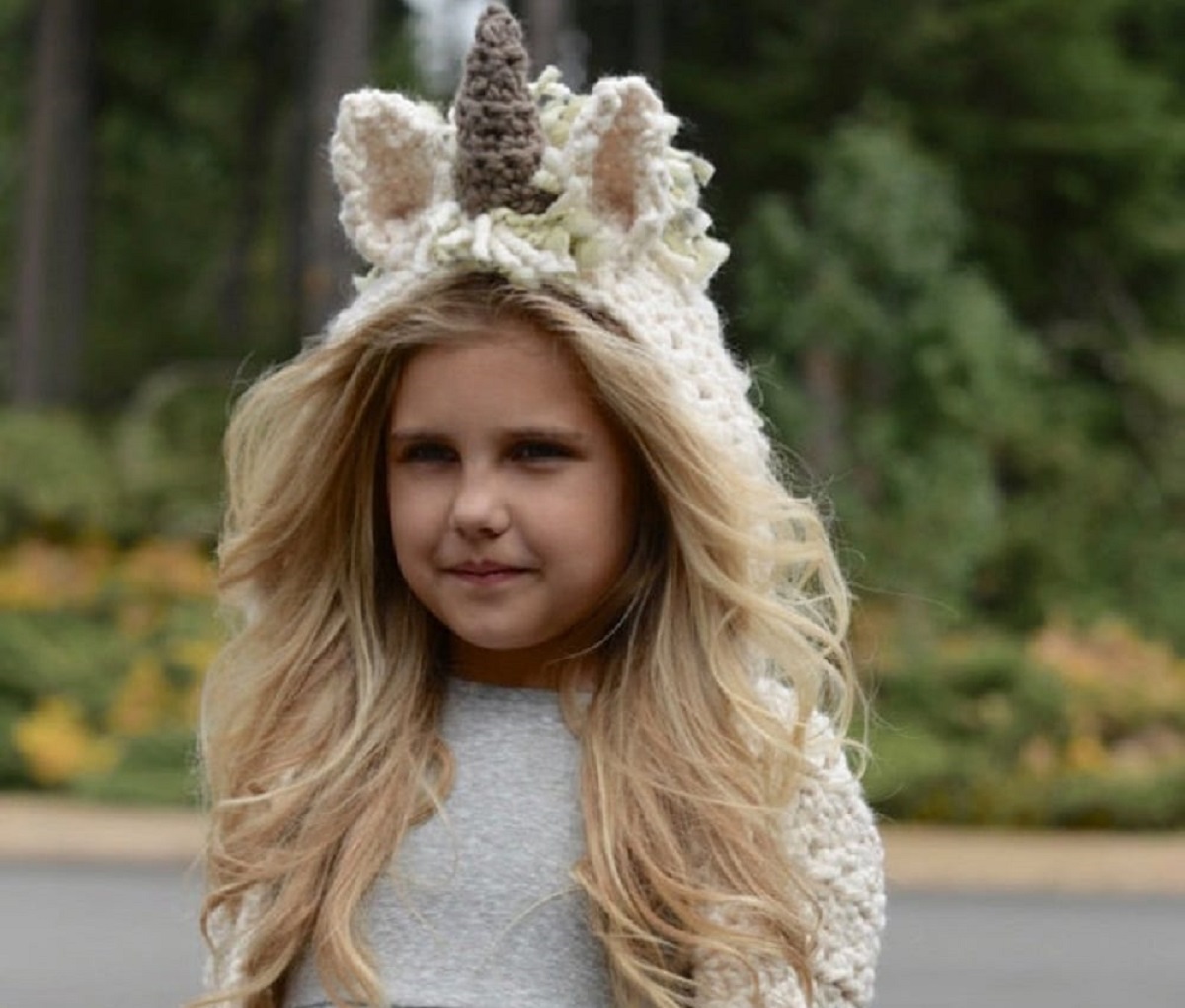 A blonde child outside wearing a unicorn hooded scarf with a grey horn in the center.