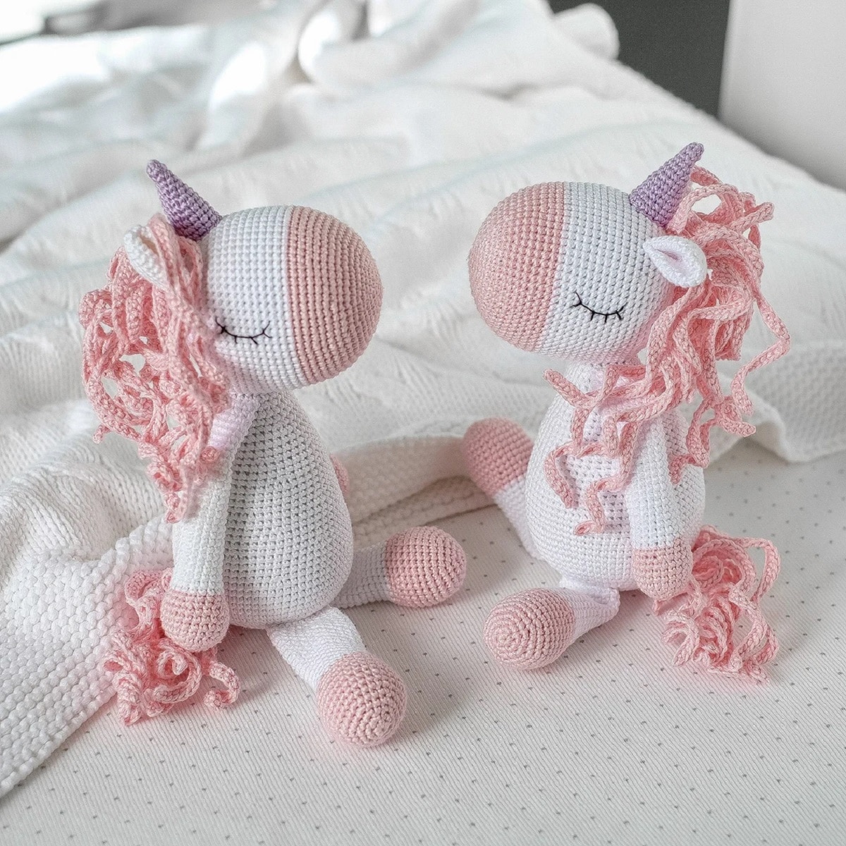 Two white crochet unicorns with pink noses, feet, curled manes, and purple horns sitting on a white blanket facing each other.