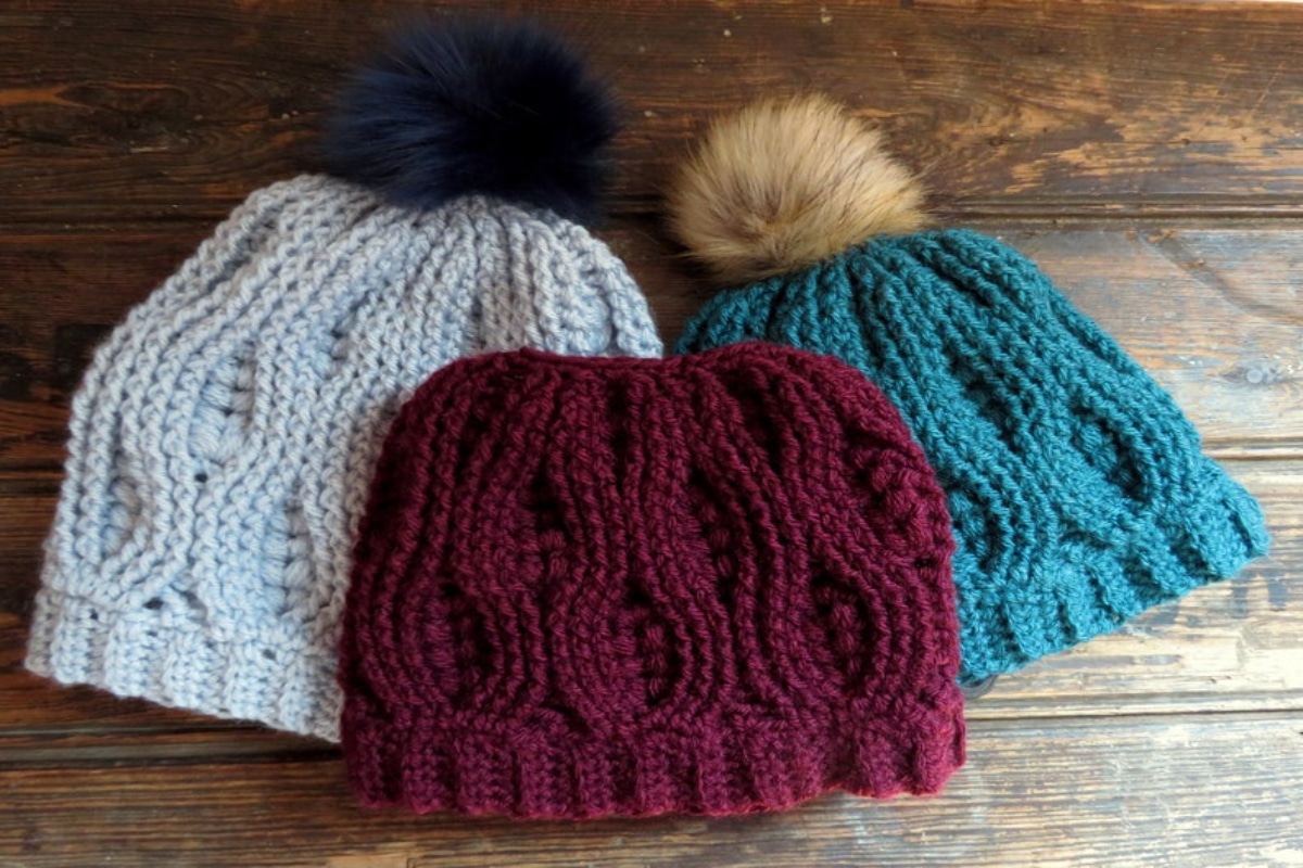 Cream, teal, and purple crochet hats with fluffy bobbles on top spread out on a dark wooden floor.