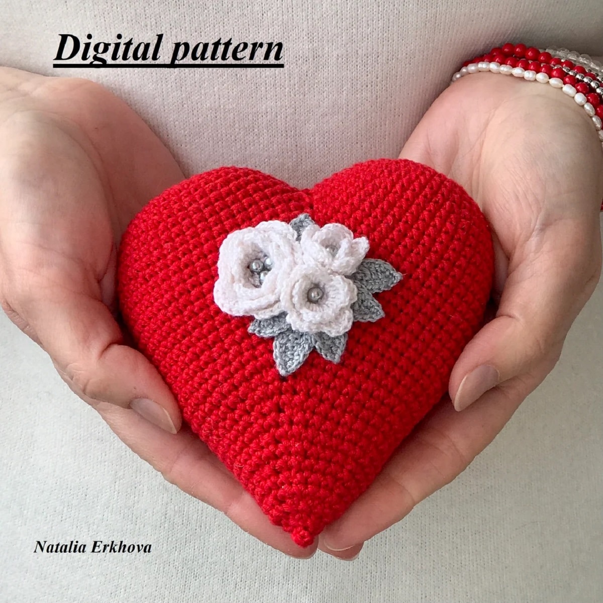 Crochet red heart stuffed with three small white flowers and green leaves stitched in the center held by two hands. 