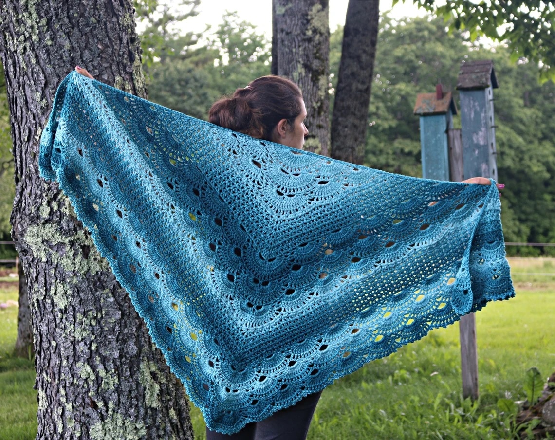 The back of a woman next to a tree with a diamond shaped crochet shawl with light and dark blue diagonal stripes. 