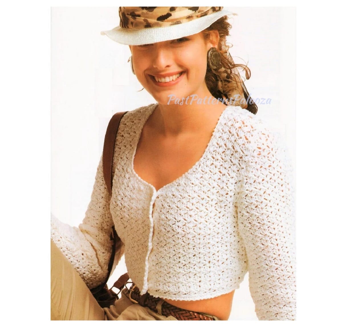 Brunette woman in a hat wearing a cream cropped lacy crochet cardigan with buttons down the front and long sleeves.