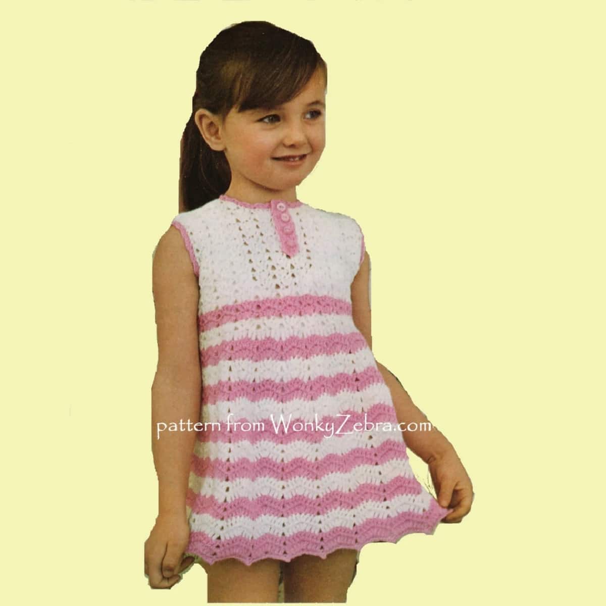 Young girl standing wearing a cream and pink crochet dress with zig-zag stripes from the chest to the bottom of the dress.
