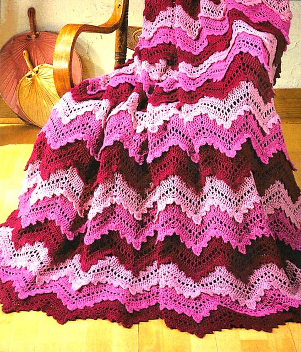 Lace style purple, pink, and red ripple crochet afghan draped over a wooden chair, falling onto wooden flooring.