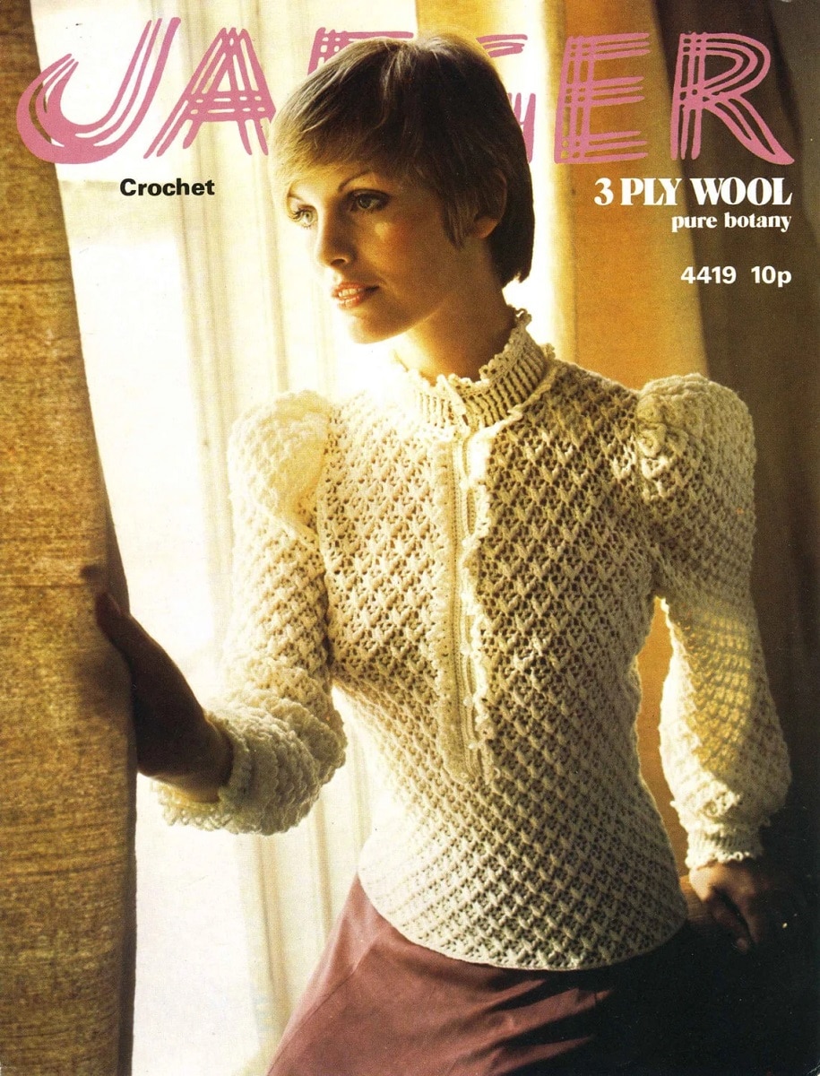 Blonde woman standing by a window in a Victorian style crochet blouse with a high neckline and a ruffle down the center.