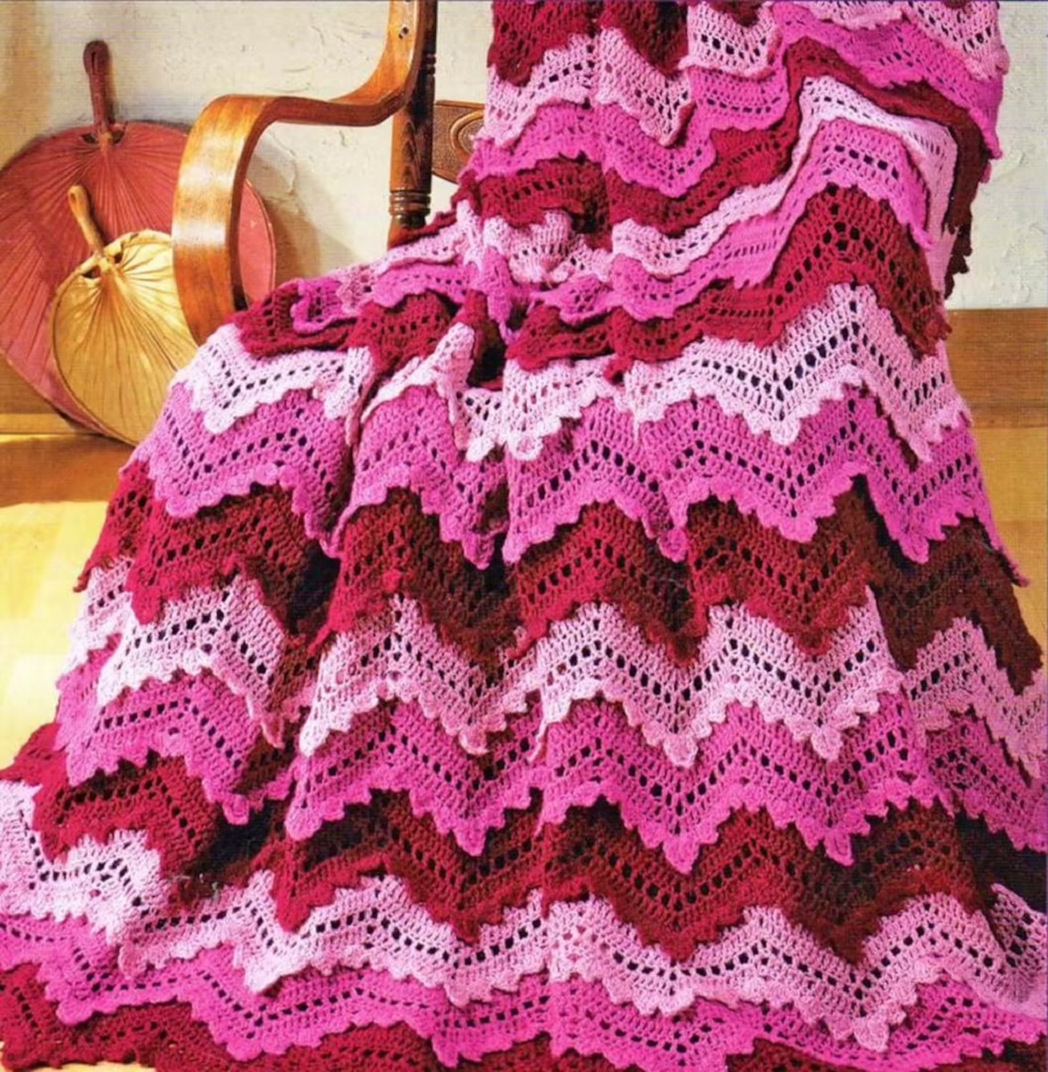 Large pale pink, pink, and purple zig zag crochet blanket draped over a wooden rocking chair.