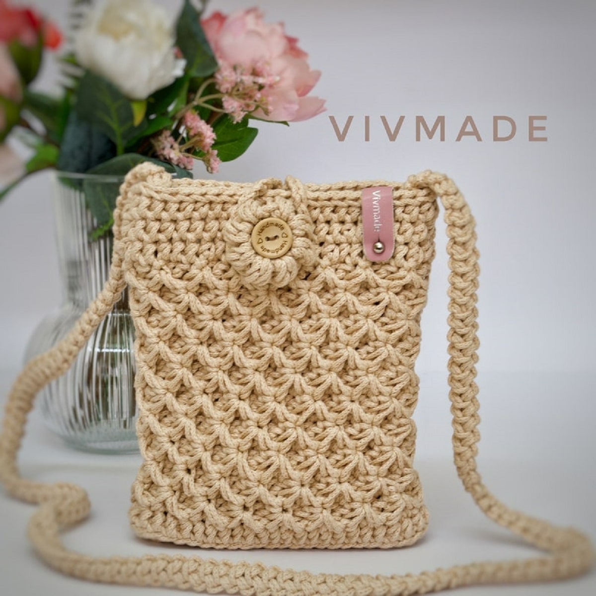 Light brown crochet bag with a fan shaped pattern, a flower at the top of the bag and a long thick braided strap. 