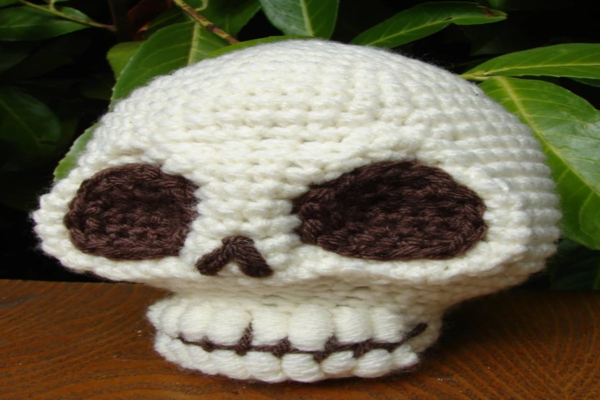 White crochet stuffed skull with brown eyes and nose and defined teeth, sitting on a dark wooden table.