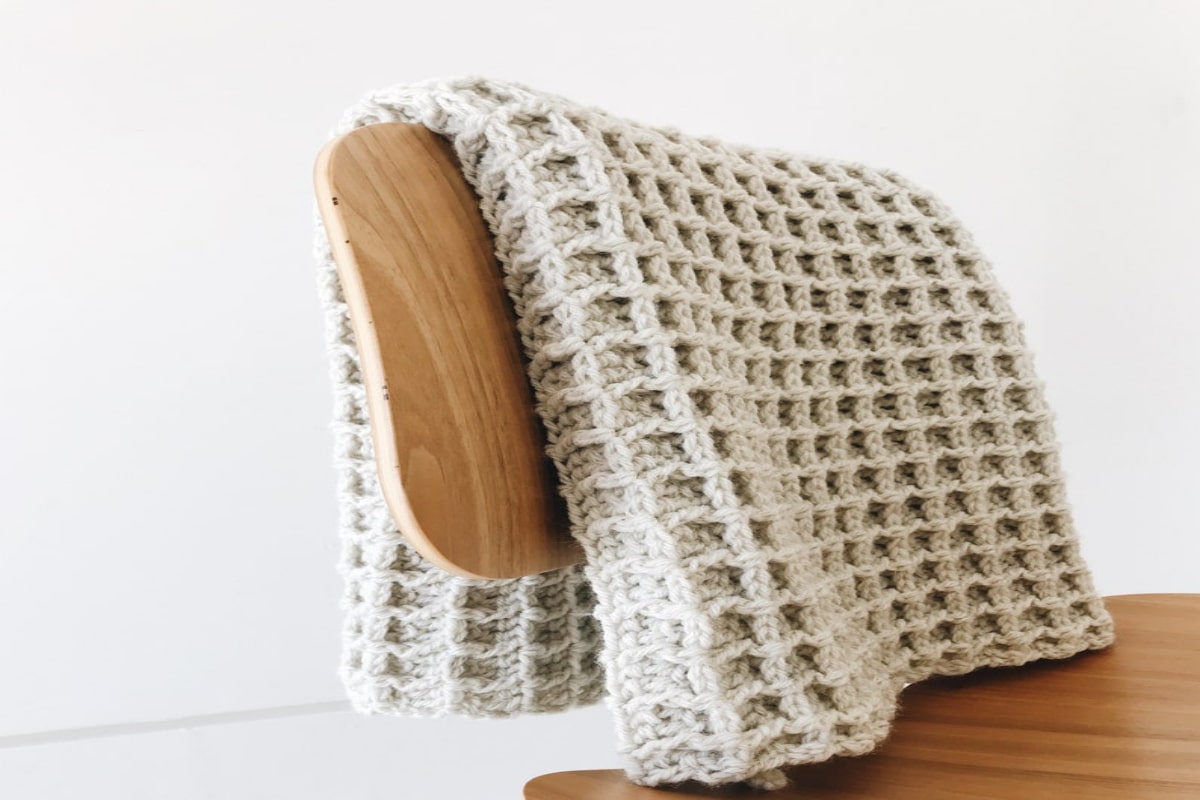 Cream waffle stitch crochet blanket folded over a wooden chair next to a wooden table.