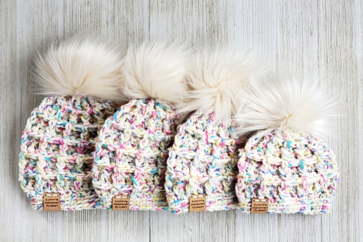 Crochet waffle stitch beanies with a white fluffy bobble using cream, blue, pink, and yellow yarn blended together. The hat is displayed in four sizes next to each other.