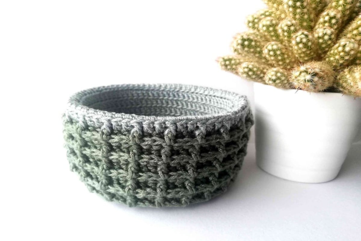 Small green waffle stitch crochet bowl with a lighter green yarn used for the inside next to a white pot with a cactus in.
