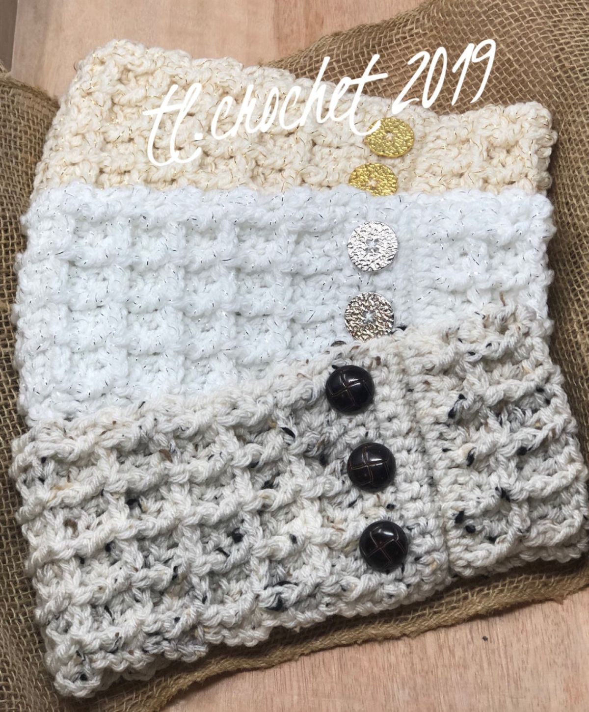 Pale gray, white, and light yellow crochet ear warmers with waffle design and three buttons off the center on a brown hessian background. 