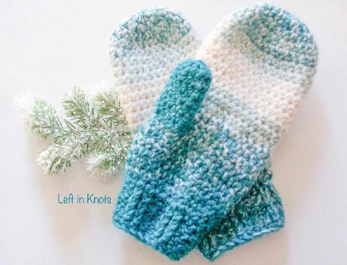 Dark blue, light blue, and white crochet mittens on top of each other with snowy leaves and a white background.