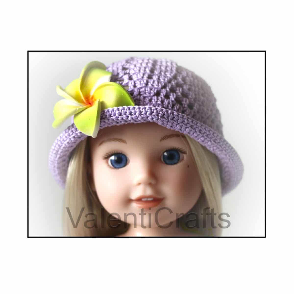Blonde doll wearing a purple crochet hat with a yellow and green lily on the side on a white background.