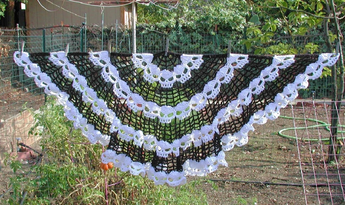 Black crochet half moon shawl with white skulls in horizontal lines hanging on a line with some grass and shrubs. 