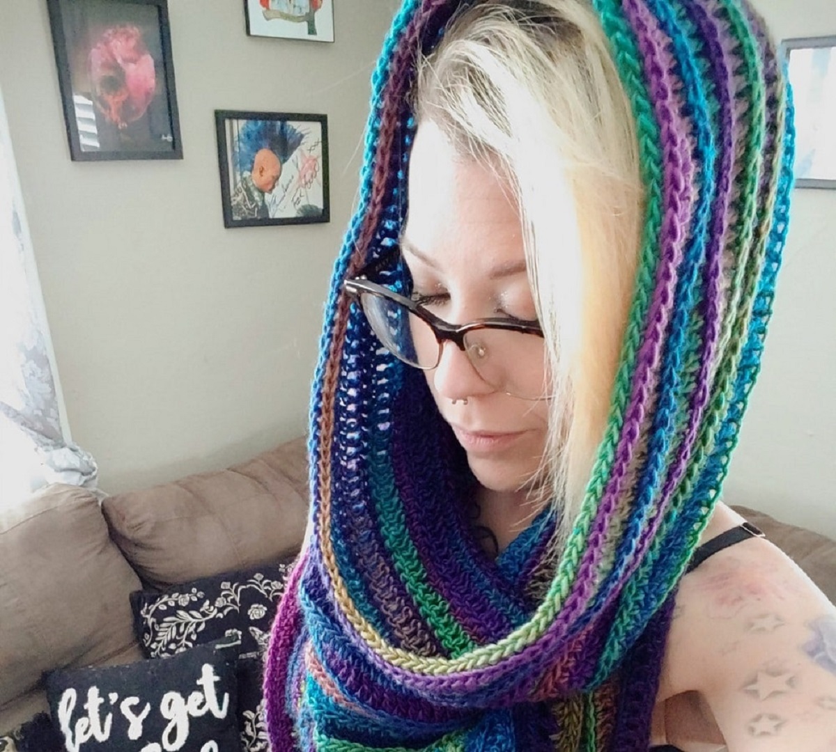 Blonde woman in glasses wearing a purple, blue, green, and yellow striped hooded scarf.