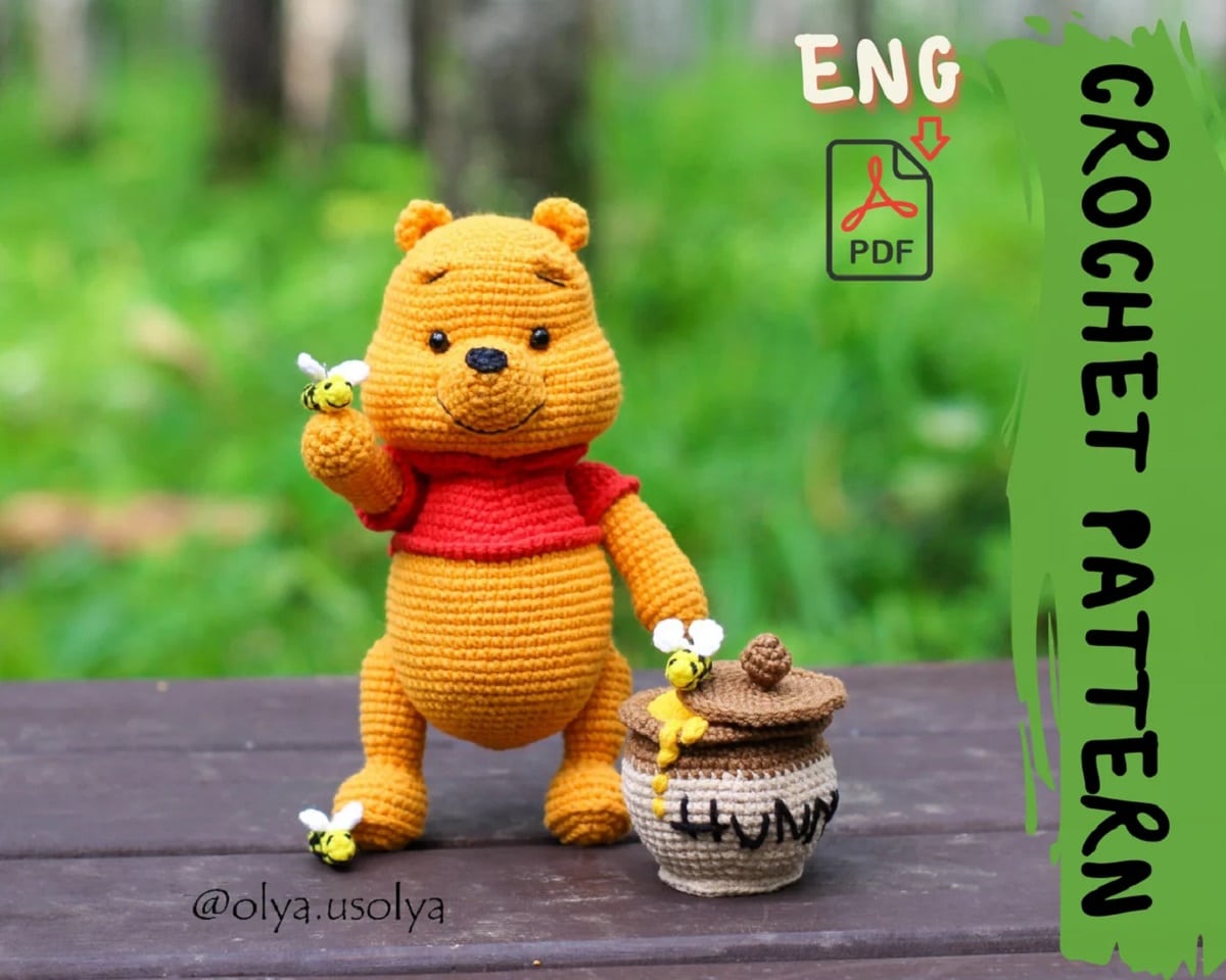 Crochet stuffed Winnie the Pooh in his classic red jumper standing next to a pot of honey and some small crochet bees.
