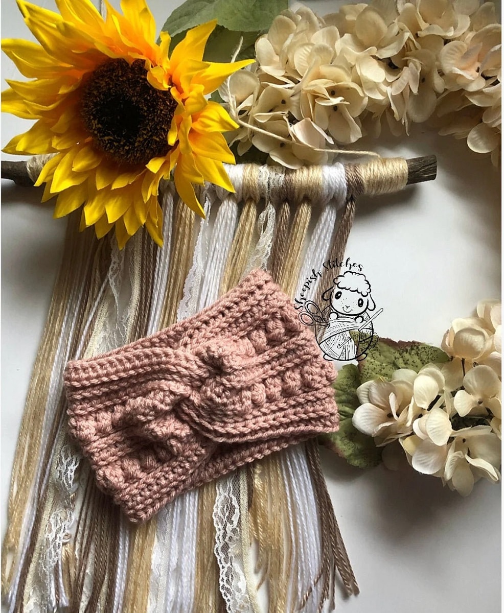 A pale pink crochet headband with twisted design at the front and vertical rows of small circles lying on different colored yarn with flowers around it.