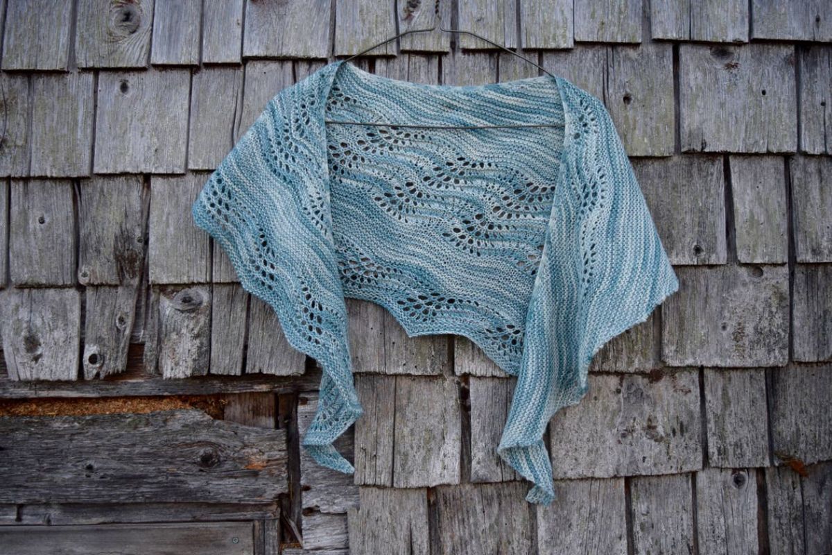 Light blue crochet shawl with a slight scalloped edge on a hanger in front of a gray background.