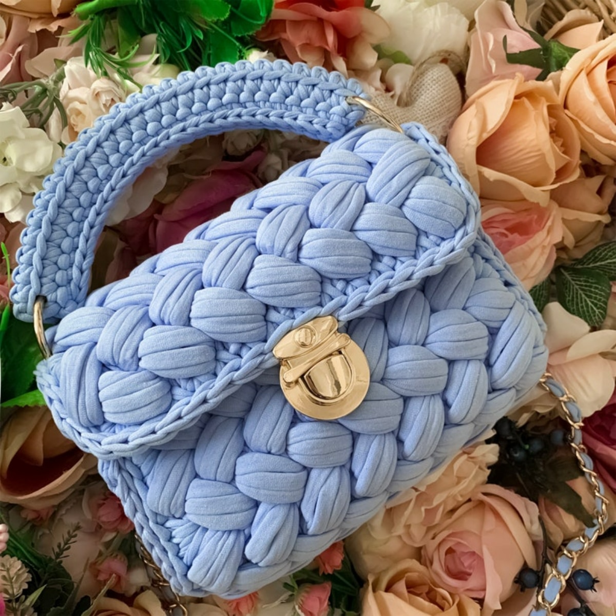 A small blue crochet handbag with a gold buckle in the center to secure it and a short thick blue braided handle on top.