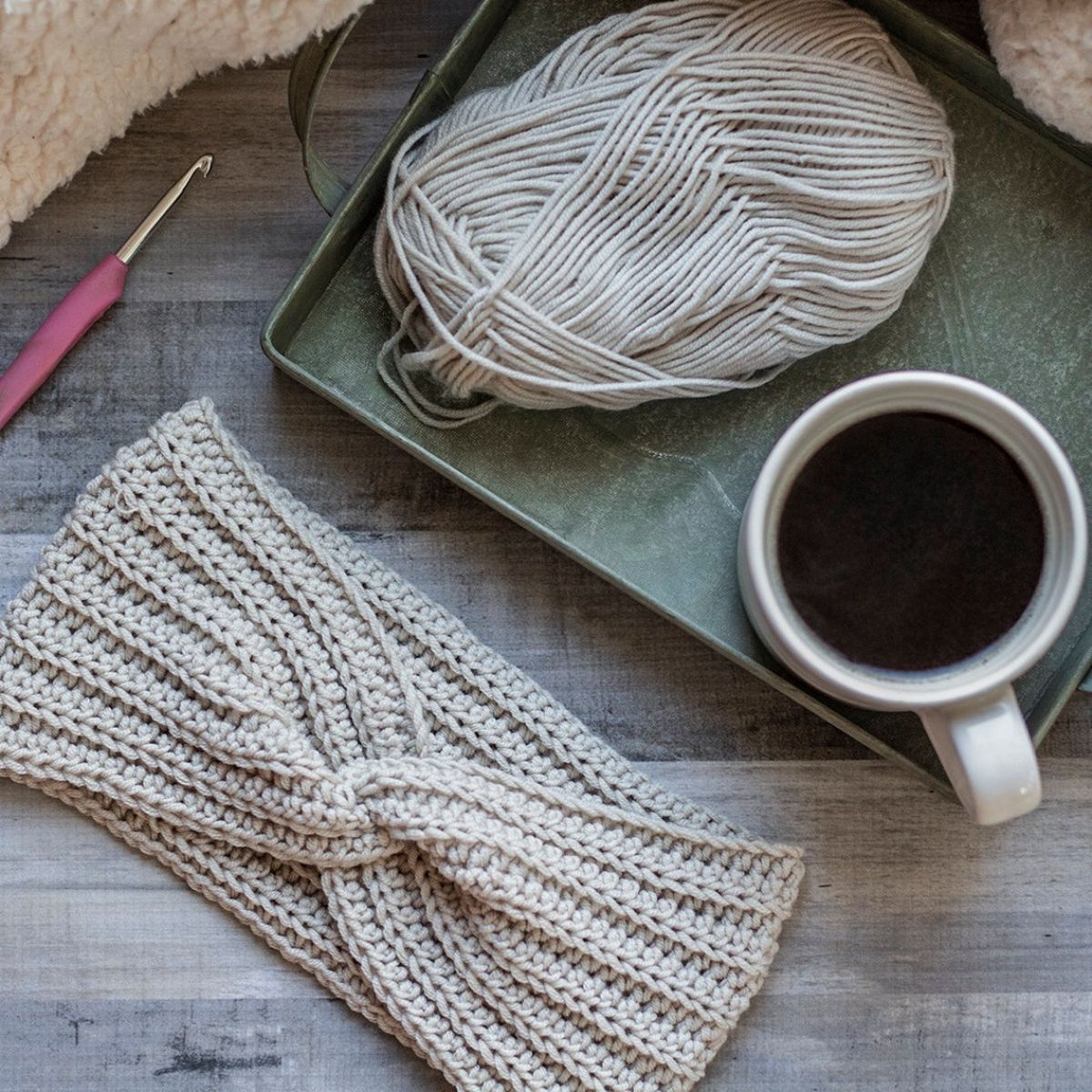 A cream crochet ear warmer with twisted design on the front on pale wooden flooring next to a cup of coffee, cream yarn, and crochet hook. 