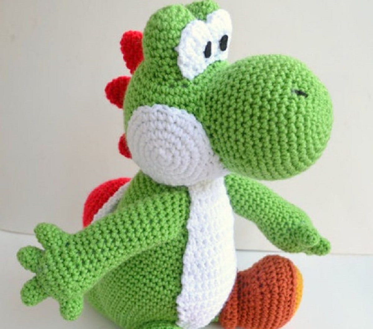 Crochet green and white Yoshi sitting sideways with one arm stretched behind him and red spikes running down his back.