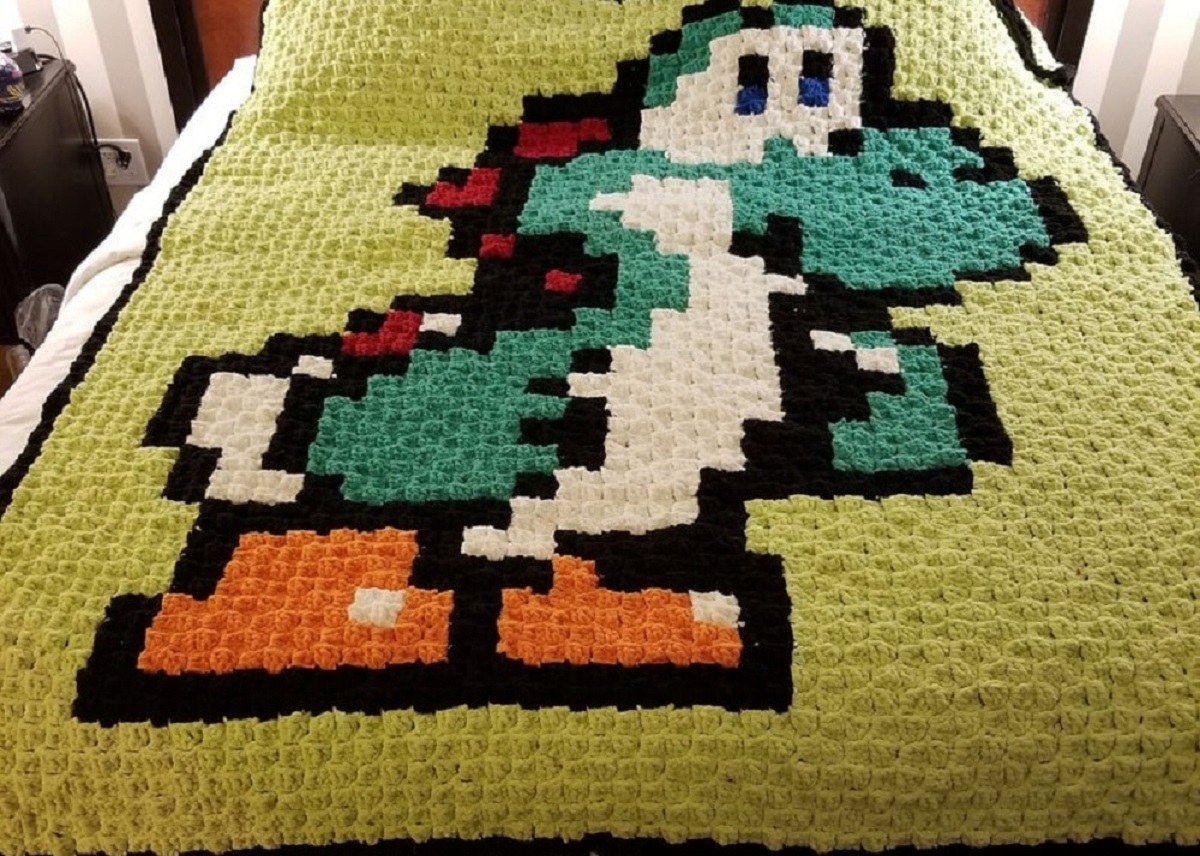 Pixel style crochet blanket with a large green and white Yoshi standing sideways so you can see his red spikes and orange feet.