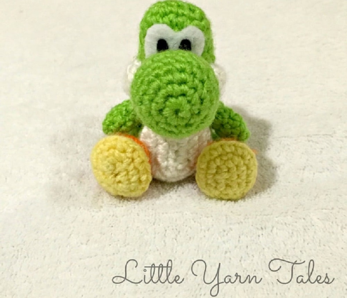 Small crochet stuffed green and white Yoshi with yellow feet sitting down on a cream carpet.
